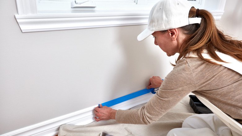 A woman tapes off baseboards with blue painter's tape before painting them.