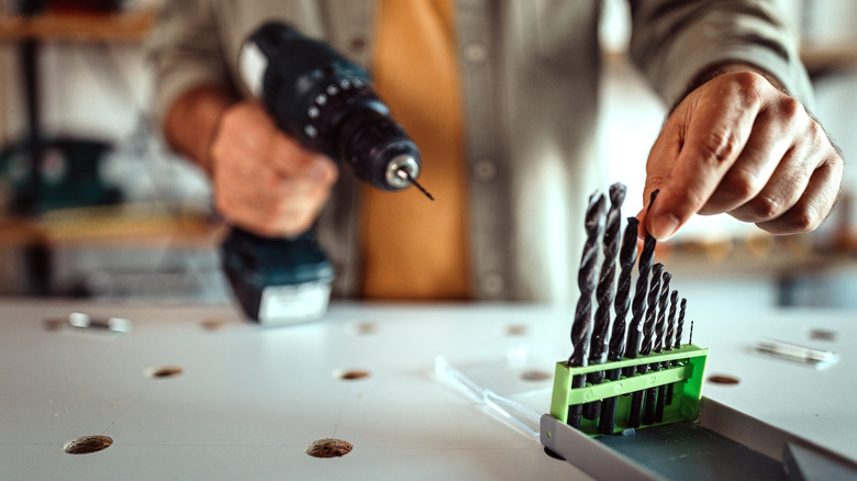 Close-up of unrecognizable craftsperson holding a hand drill and picking a drill bit from a set on a worktable.
