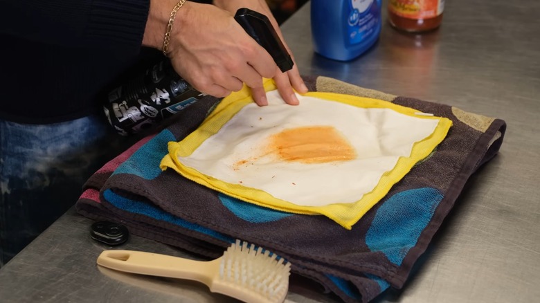 Hands spraying a stain-removing spray on an orange mark on a white cloth