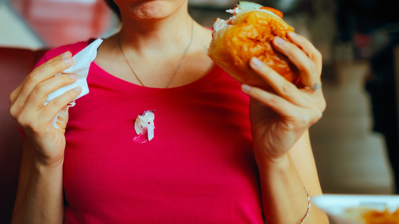 Woman holding a burger in one hand and a napkin in other to remove mayonnaise stain on her red shirt