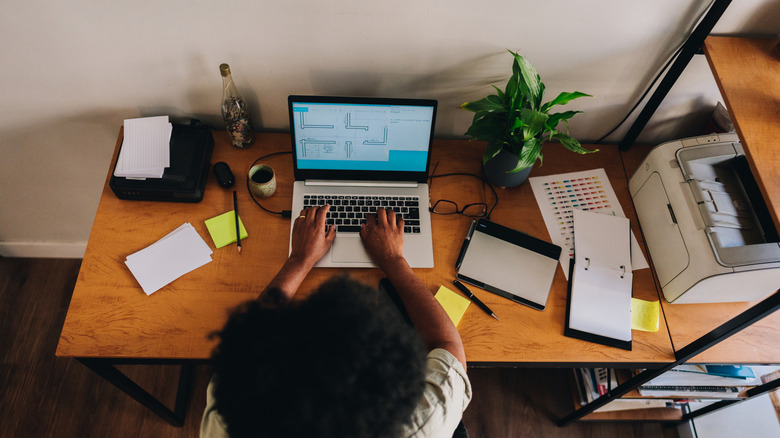 A person works at an at-home desk with a printer on a shelf near them