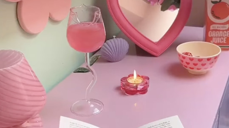 A rose-shaped wine glass filled with pink liquid on a pink desk with other decor