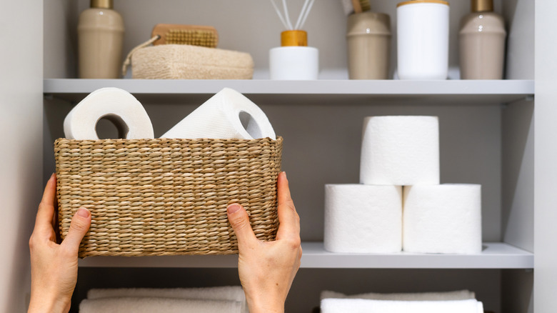 A pair of hands placing a wicker basket with two rolls of toilet paper in it on a bathroom shelf