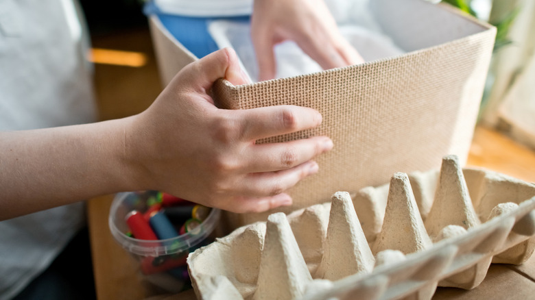 A person sorts out craft supplies with an opened cardboard egg carton on the tabletop.