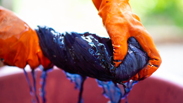 A person wearing orange gloves is twisting a freshly-dyed piece of purple fabric over a bucket