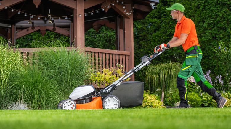 A person in an orange shirt and green trousers mowing the lawn in front of a wooden pagoda in a garden with lush and vibrant plants.