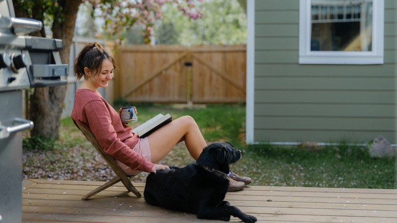 A woman in a long sleeve red shirt drinking out of a mug next to a black dog. Wooden and white picket fences are visible in the background