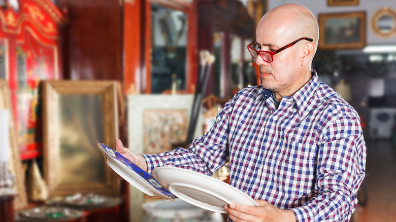 A man wearing red glasses examines two china plates in an antique store.