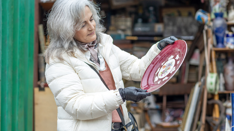 Woman with silver hair and white puffy coat holds a colorful red ceramic plate with gloved hands at a flea market or bazaar outside