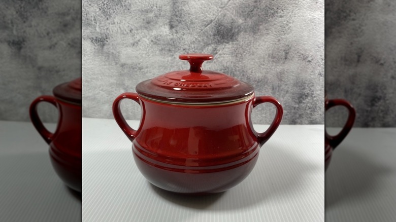 A red Le Creuset bean pot displayed against a white and gray background.