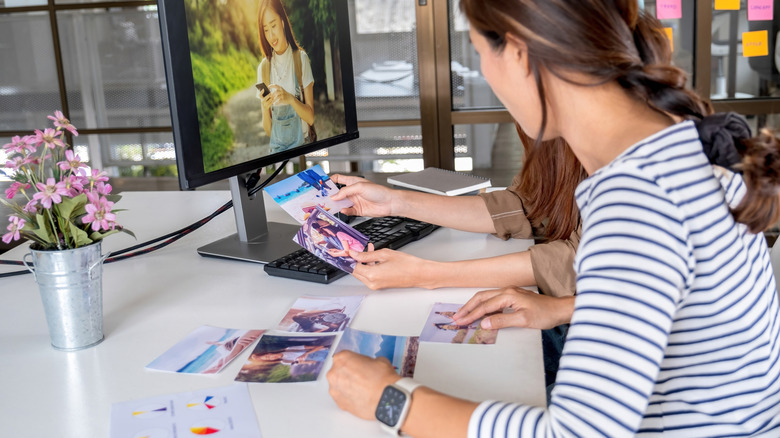 Two people sitting at a desk with a computer and a vase of flowers sorting through some photos