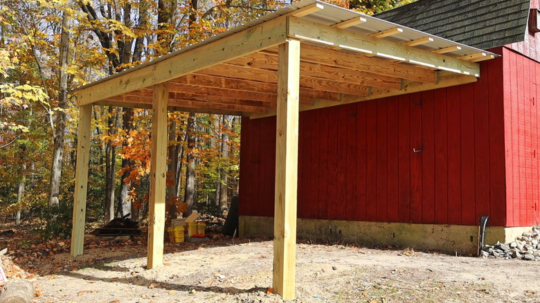 A lean-to shed with no walls built on the side of a red barn near trees with orange and yellow leaves