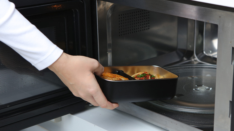 A hand placing a black and silver container with food into the microawave