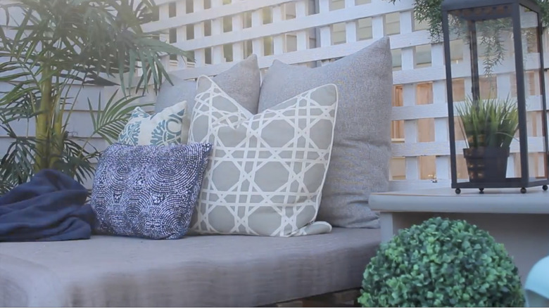 A DIY wood pallet outdoor daybed is covered in grey cushions and grey, white, and blue pillows.