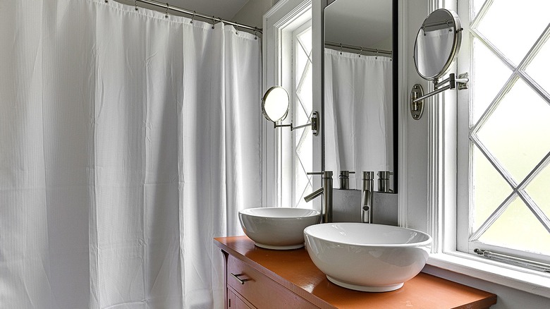 A bathroom vanity with two vessel sinks beside two windows and a white shower curtain