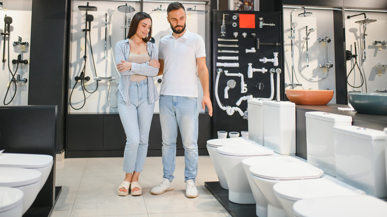 Man and woman deciding on which toilet to buy in store while man sits on one and the woman crouches next to him