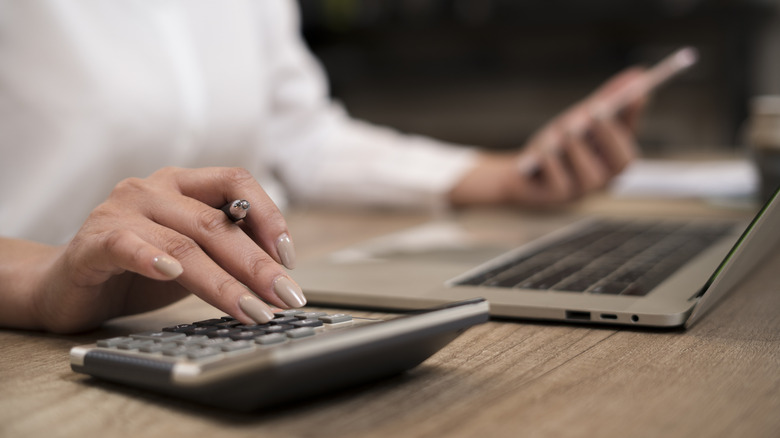 Person in a white shirt sitting at a wooden desk using calculator while referring to phone and laptop