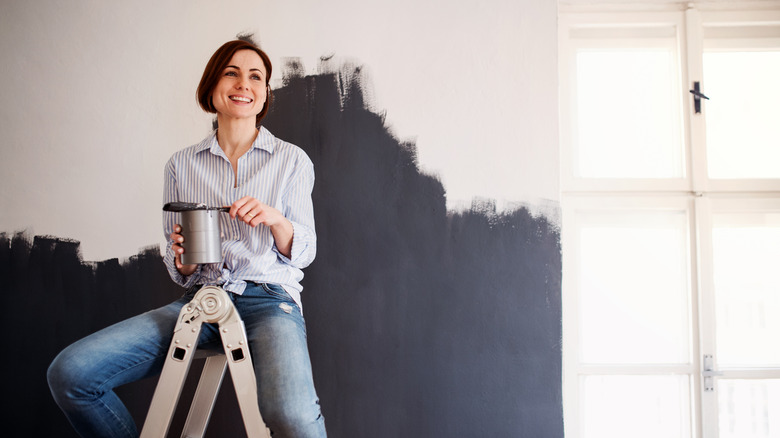 A light-skinned woman with short, dark hair smiles while taking a break from painting a wall a dark charcoal color.