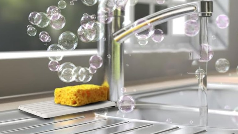 A yellow sponge sitting on a NiuYichee Diatomite Coaster next to a kitchen sink with bubbles in the air