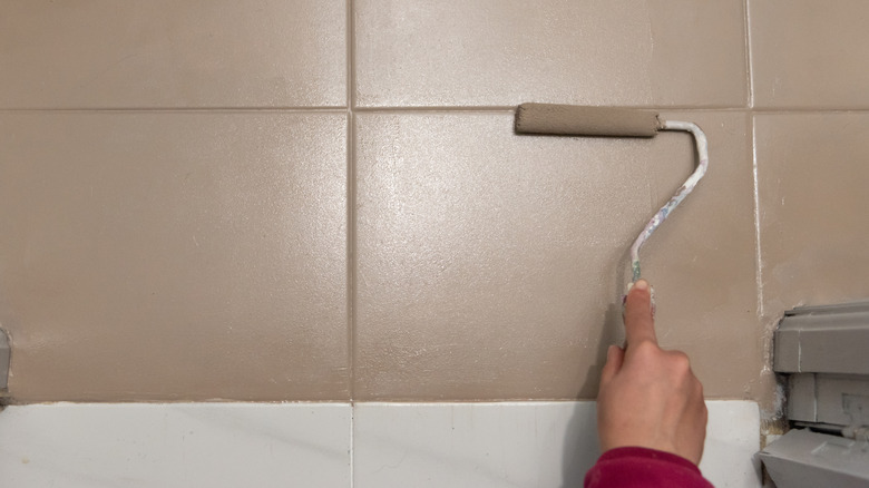 Someone painting over wall tiles with a roller and beige paint