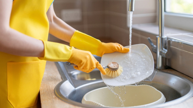 Woman in a yellow apron and gloves washing dishes with a scrub brush