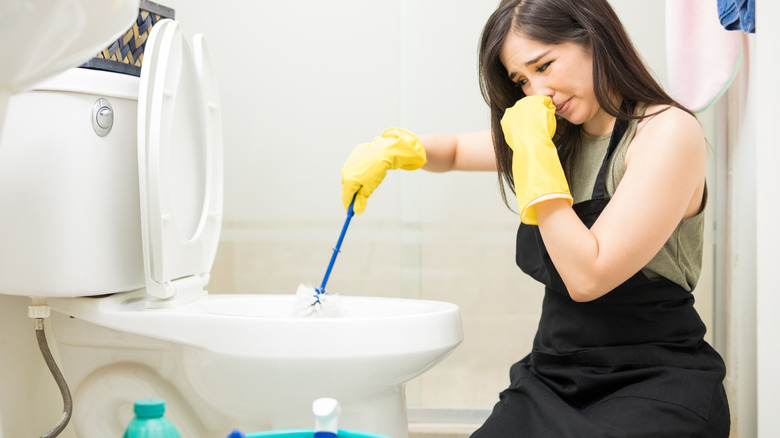 A dark-haired woman holds her nose while cleaning a dirty toilet