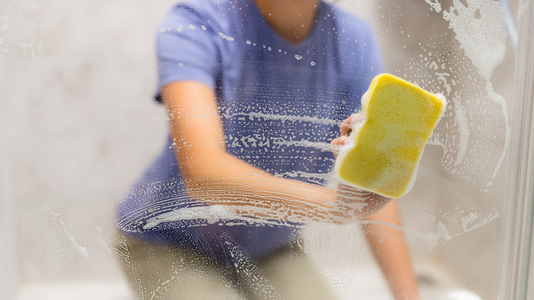 A person kneels while using a yellow sponge to clean glass shower doors
