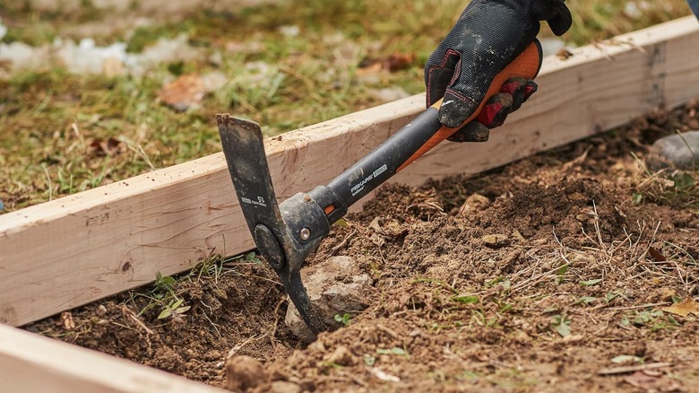 A gloved hang digs into a lumber-framed section of earth with a Fiskars pickaxe.