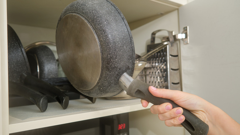 A person puts a frying pan into an overstuffed upper cabinet.