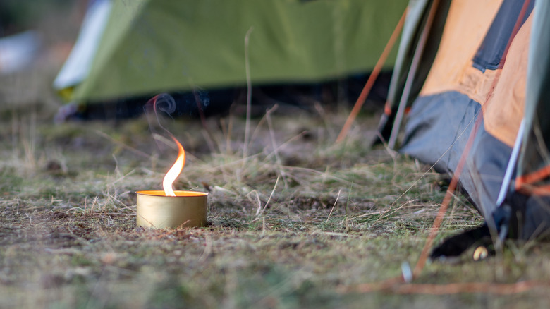 A lit citronella candle in a small metal container, sitting on the ground next to a tent