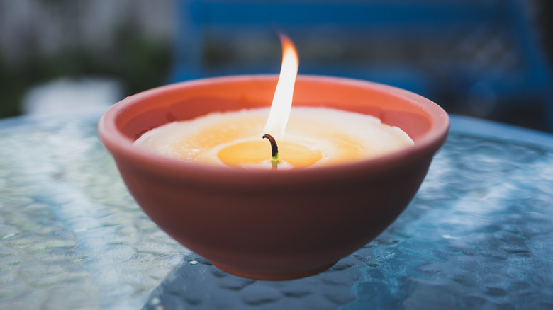 A lit citronella candle in a reddish brown holder burning on a glass patio table