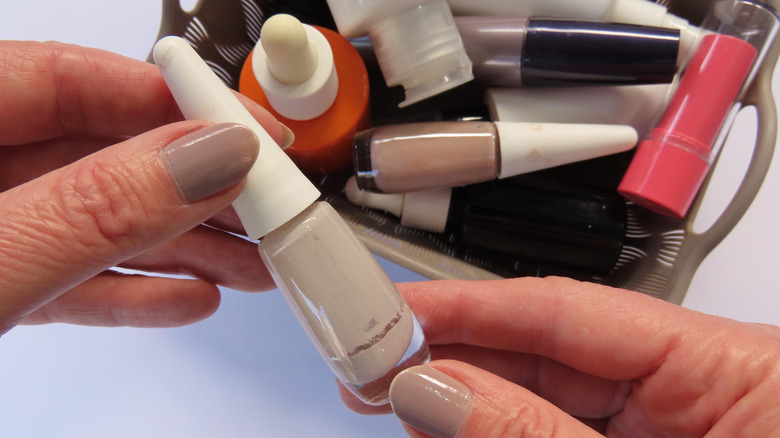 Two hands hold a bottle of nail polish above a basket containing more polish and other makeup.
