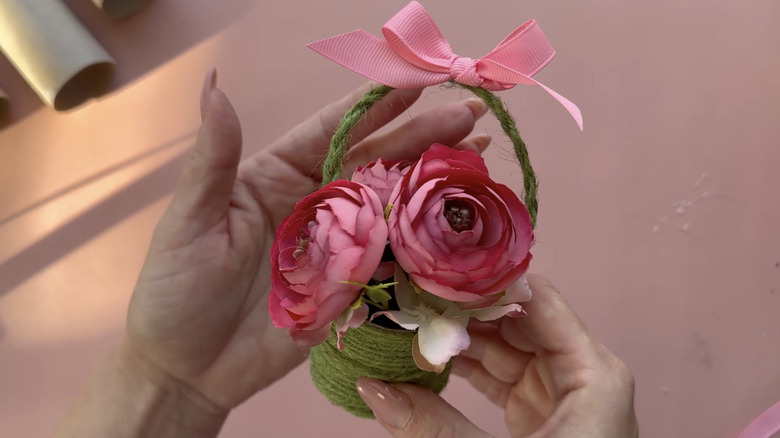 A mini flower basket made from paper towel tubes with artificial pink peonies inside and a pink bow on the handle.