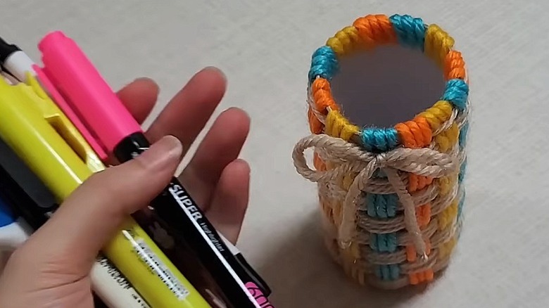 A person holds pens and markers in one hand next to a yarn-covered paper towel storage cup