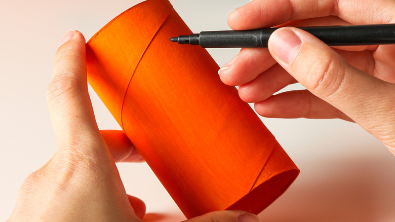 Hands drawing with pen on an orange-painted toilet paper roll tube.