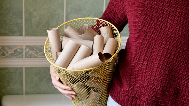 A person holds a yellow wastepaper basket full of empty toilet paper rolls.