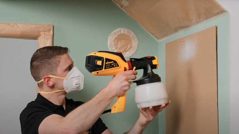 Young man using corded paint sprayer on indoor wall