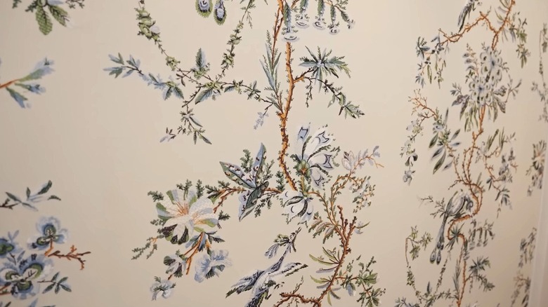 Close-up of embroidered wallpaper with a cream background and nature inspired designs in green, brown, and blue.