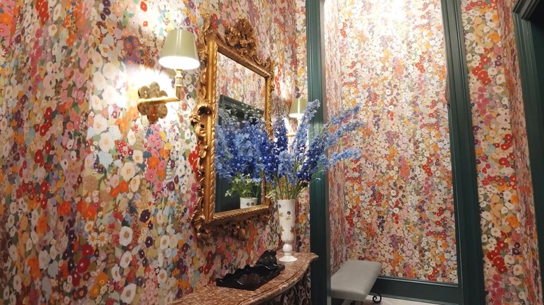 A hall is clad in colorful floral wallpaper. There is a vase with blue flowers over a floating marble shelf, as well as a gold mirror and simple sconce.