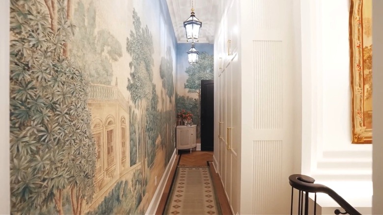 A narrow hallway with a hand painted mural of a scenic garden on the left wall. On the floor there's a runner with a geometric pattern.