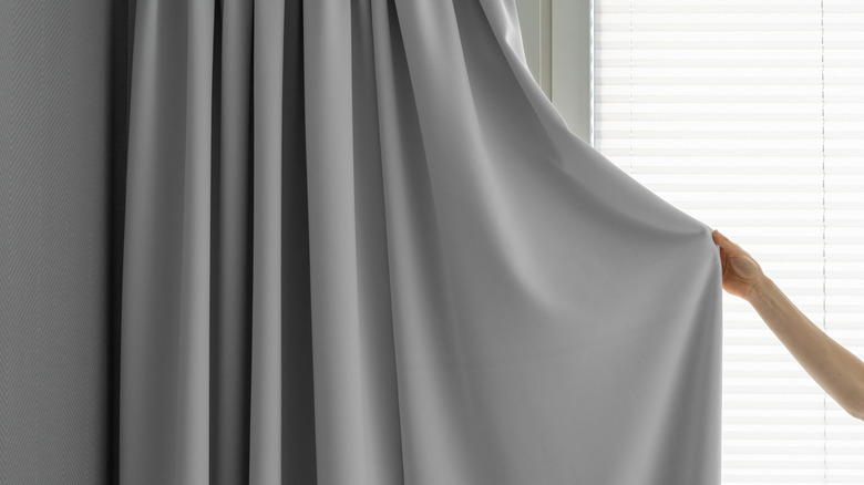 A woman grips a plain gray curtain in front of a window