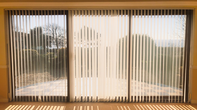 Sliding glass doors with vertical blinds, through which you can see a sunny backyard