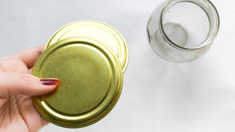 A hand with painted nails holds a lid over a jar on a gray surface.