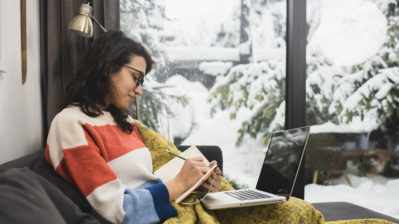 A person writes in a notebook while sitting inside under a blanket and a laptop with a winter scene out the window.