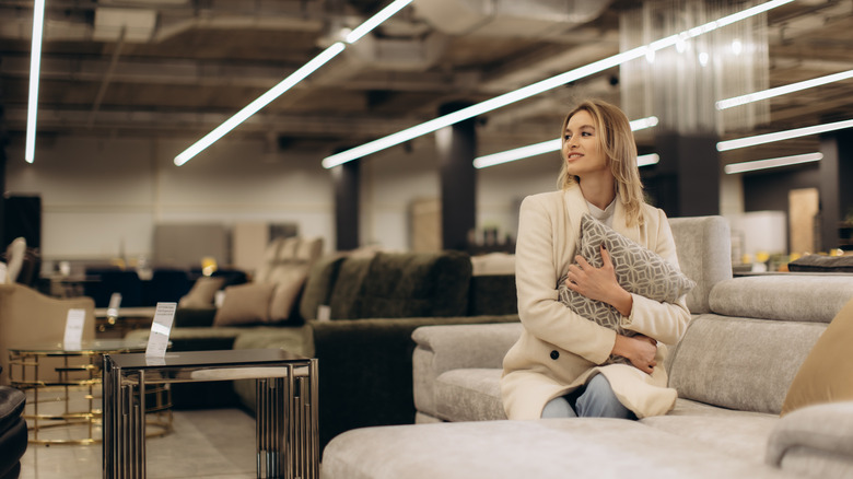 A young woman embraces a patterned pillow while seated among modern furniture in a well-lit showroom.