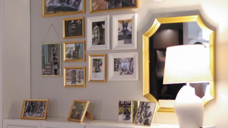 A stylish interior display featuring a gallery wall with various framed photographs in gold and white frames, a decorative mirror, and a modern lamp.
