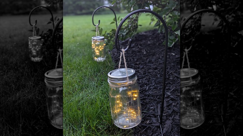 Mason jars with twinkle lights inside hanging as lanterns in a yard.