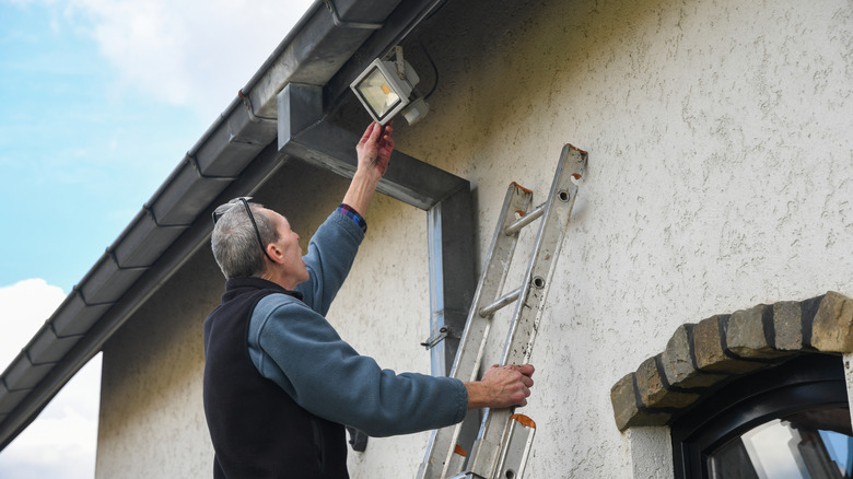 Man on ladder installing a large floodlight on the side of a home
