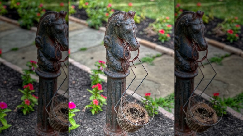 An iron horse head hitching post in a flower bed with a basket of straw attached to it
