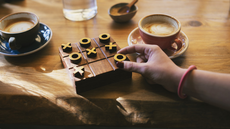 A hand making a move on a wooden tic-tac-toe game that's sitting on a wooden table and surrounded by two cups of coffee